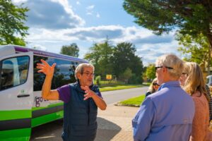 Guide talks to guests avidly on a tour in front of the bus. The sun is shining.